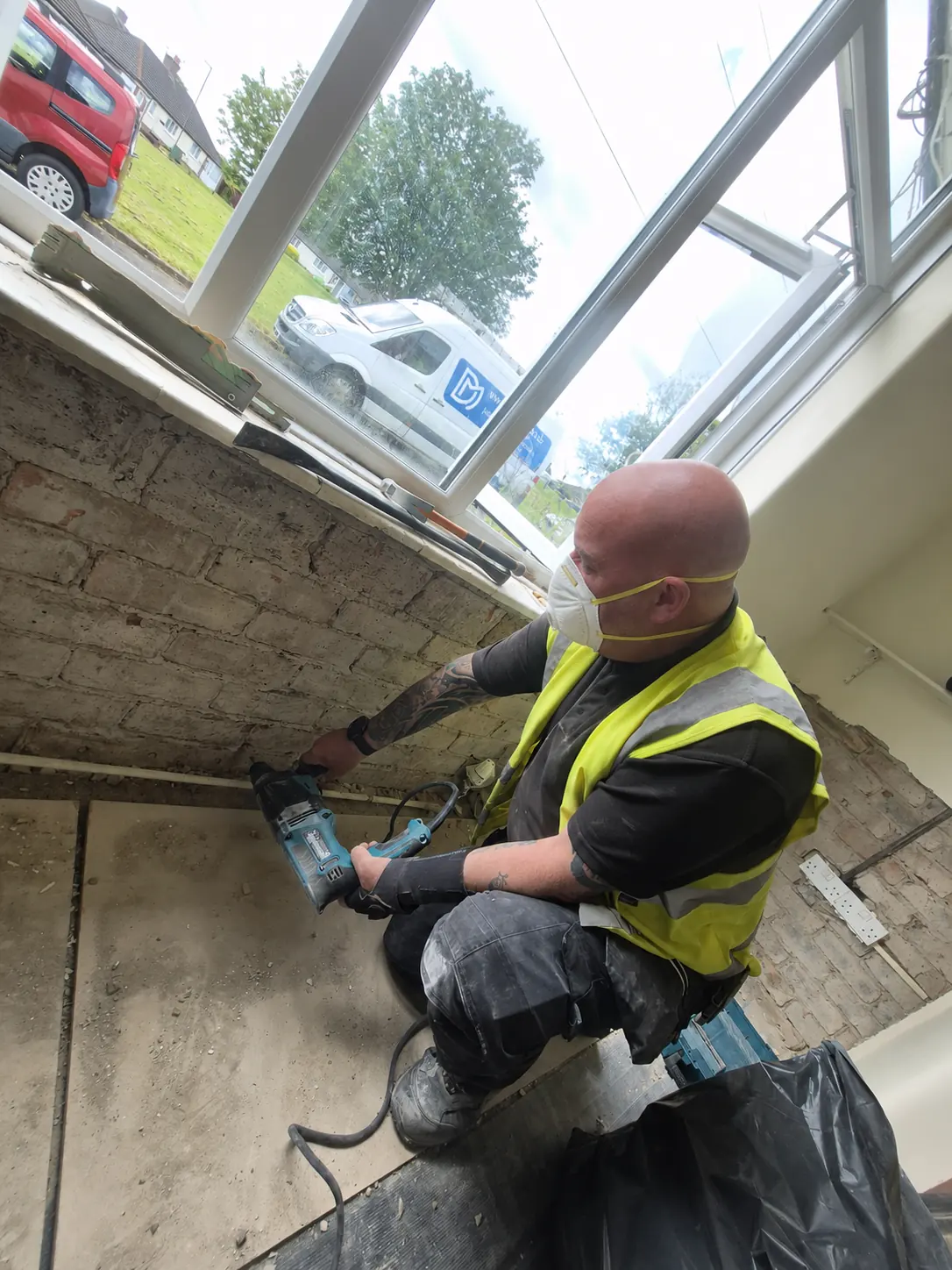 A damp proofing installer in hi-vis and a dust mask injecting damp proofing cream into drilled holes along an exposed brick wall