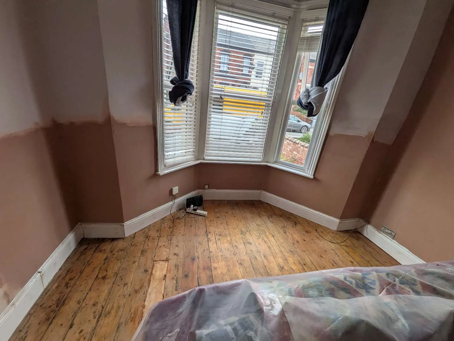 A period bay window room after damp proofing, replastered in fresh plaster up to the old line with the floorboards swept and the furniture still sheeted.