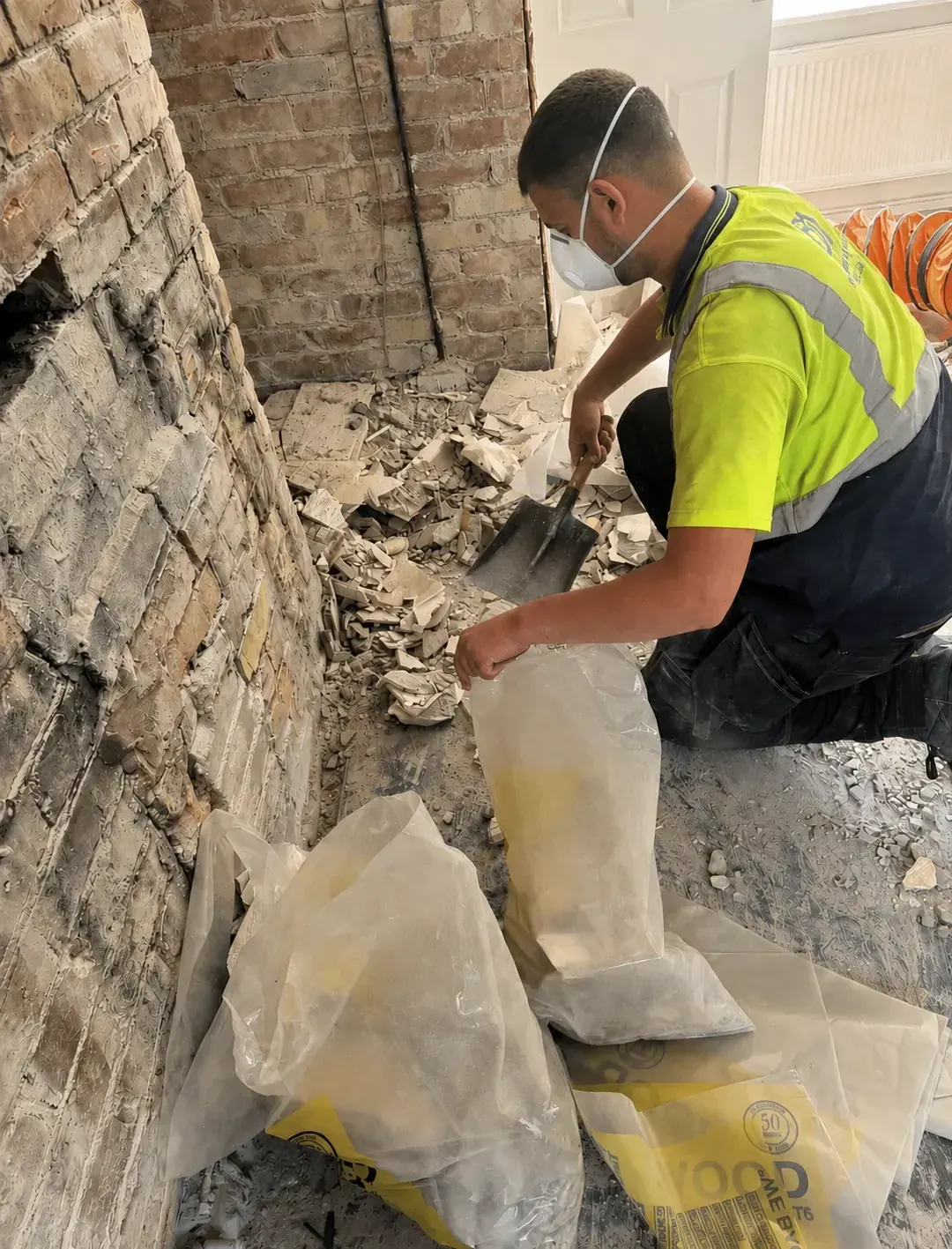 Operative in a hi-vis top and dust mask clearing salt damaged plaster into waste bags, with the wall behind him stripped back to bare brickwork.
