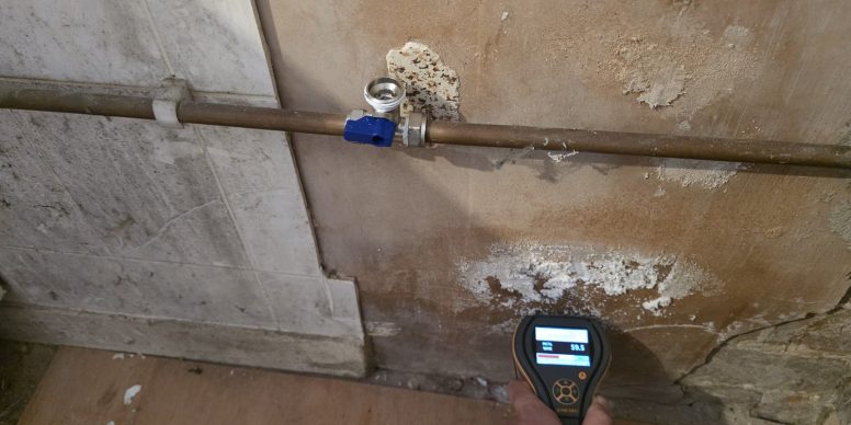White salt deposits and blown plaster spreading across a wall around a pipe, with a moisture meter on the floor below.
